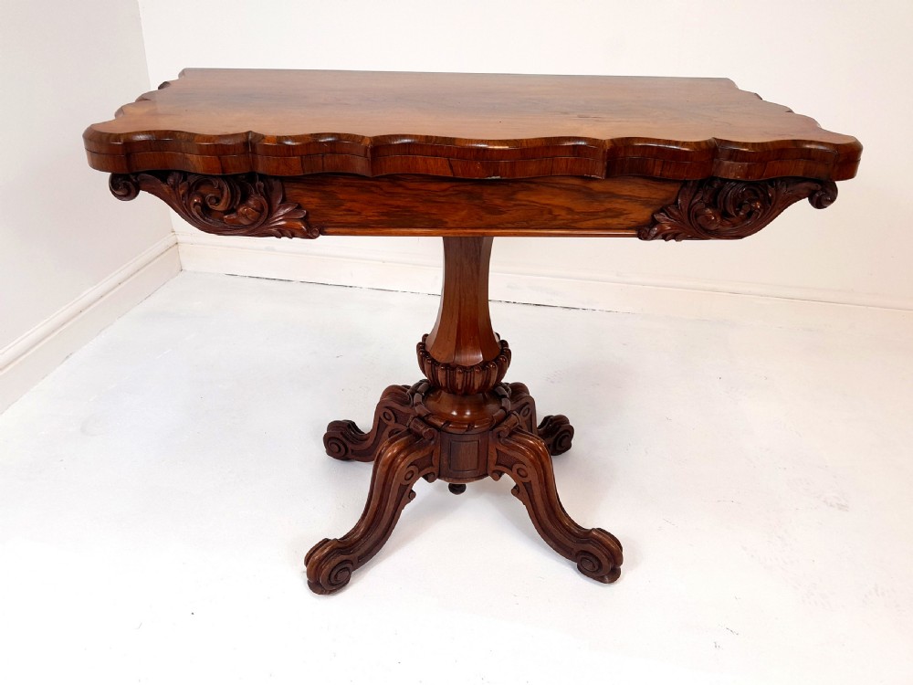 An antique Victorian rosewood card table featuring a serpentine shaped top, carved scroll frieze, central turned pedestal, and a carved quadform base. View II