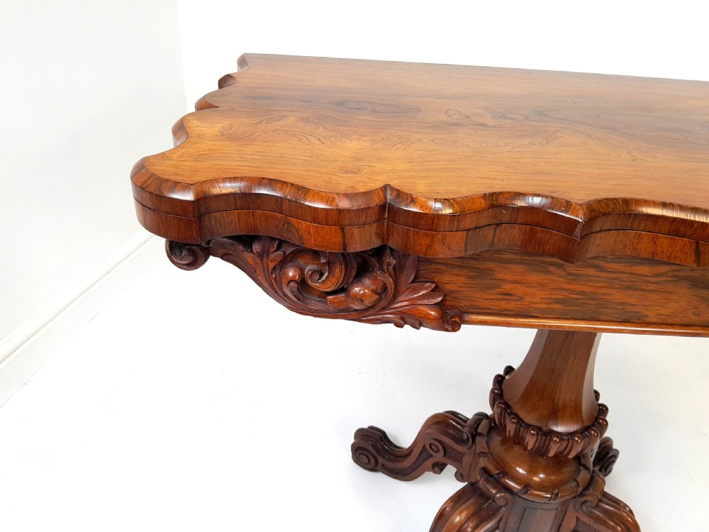 An antique Victorian rosewood card table featuring a serpentine shaped top, carved scroll frieze, central turned pedestal, and a carved quadform base. View III