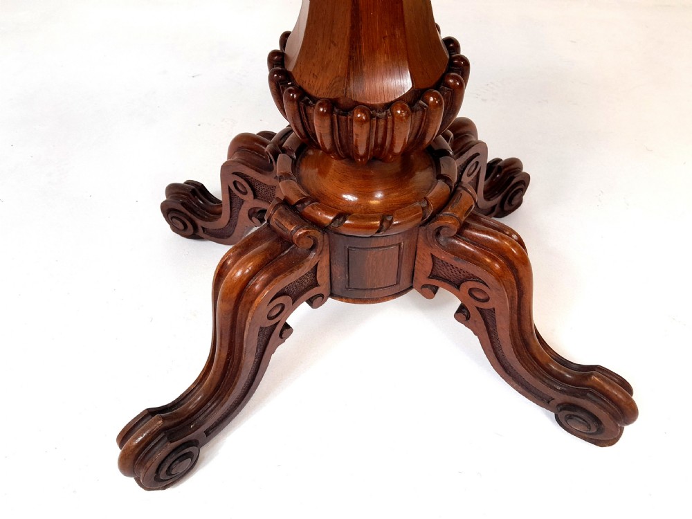 An antique Victorian rosewood card table featuring a serpentine shaped top, carved scroll frieze, central turned pedestal, and a carved quadform base. View IV