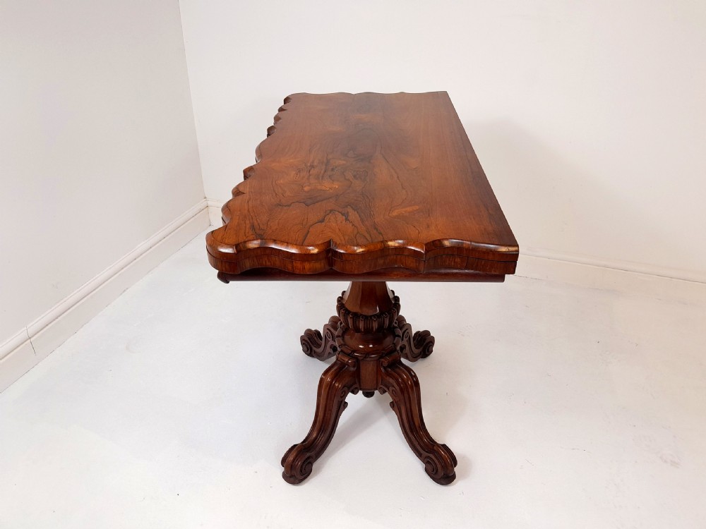 An antique Victorian rosewood card table featuring a serpentine shaped top, carved scroll frieze, central turned pedestal, and a carved quadform base. View V