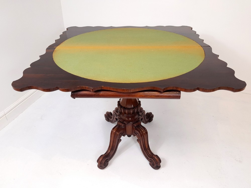 An antique Victorian rosewood card table featuring a serpentine shaped top, carved scroll frieze, central turned pedestal, and a carved quadform base. View VI