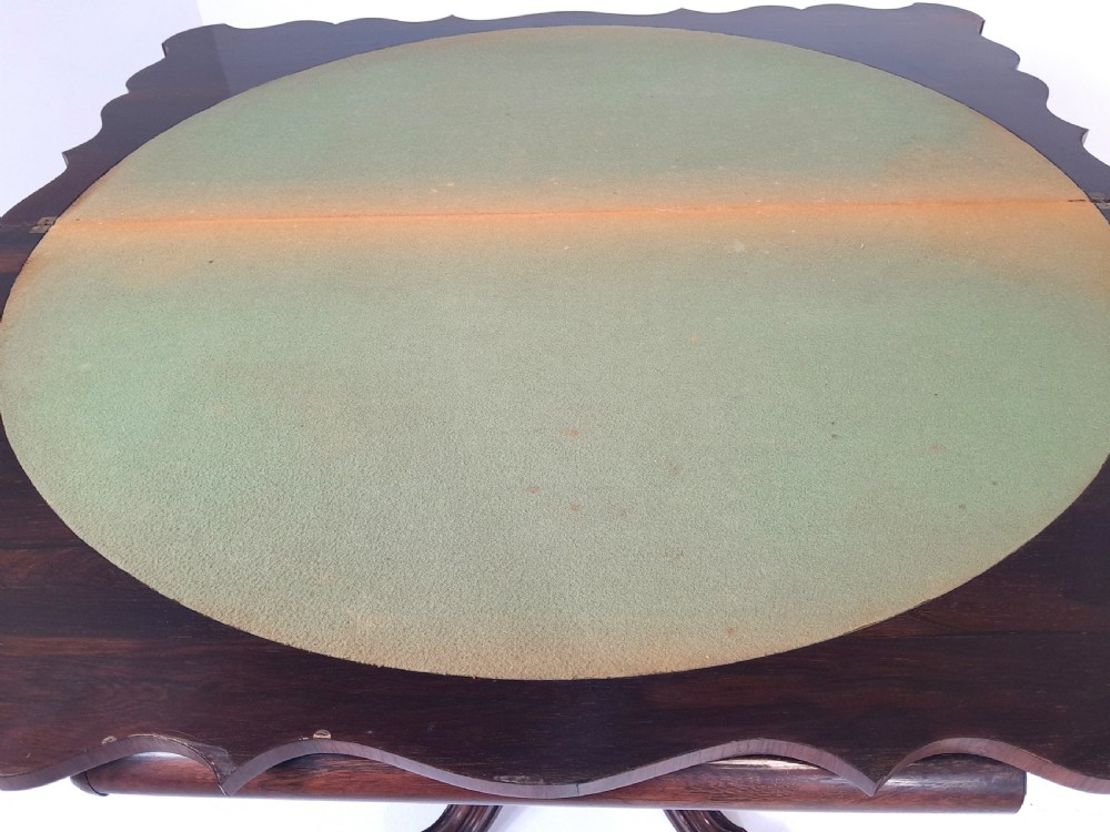 An antique Victorian rosewood card table featuring a serpentine shaped top, carved scroll frieze, central turned pedestal, and a carved quadform base. View VII