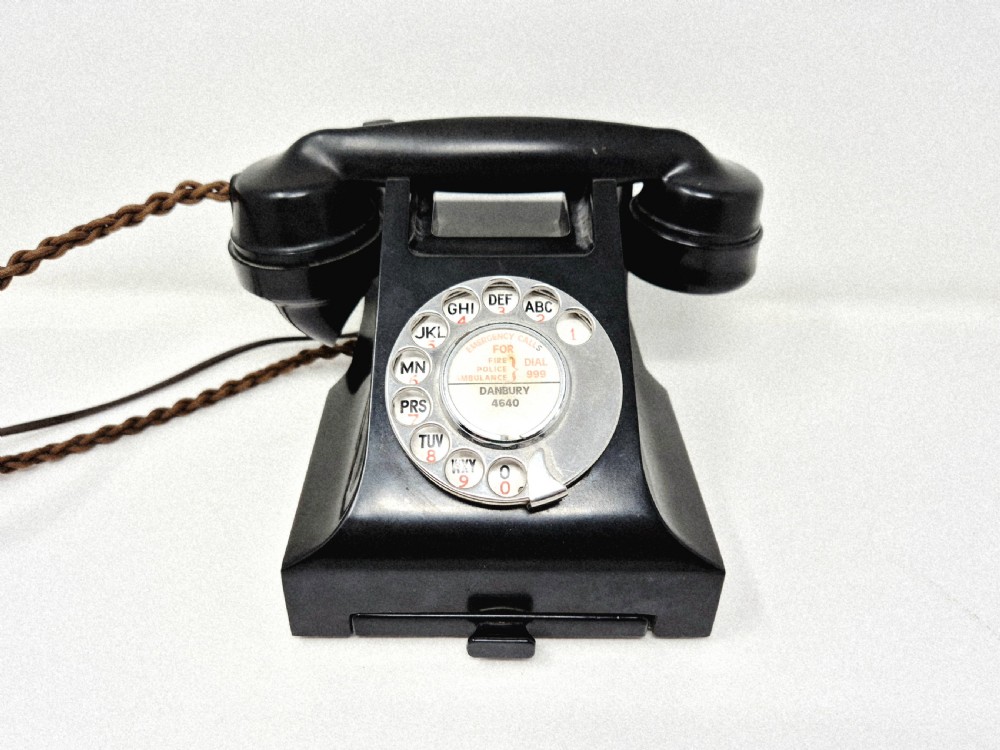 A black Bakelite rotary telephone featuring a chrome dial face, pull-out directory tray, and a braided textile cord. View III