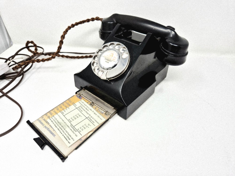 A black Bakelite rotary telephone featuring a chrome dial face, pull-out directory tray, and a braided textile cord. View IV