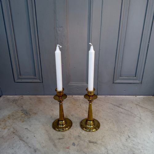 fig 1. A pair of Arts and Crafts brass candlesticks featuring tapered stems, circular drip pans, and stepped circular bases holding white taper candles.