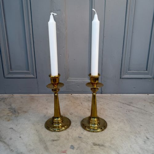 fig 2. A pair of Arts and Crafts brass candlesticks featuring tapered stems, circular drip pans, and stepped circular bases holding white taper candles.