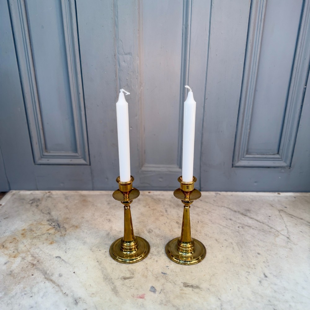 A pair of Arts and Crafts brass candlesticks featuring tapered stems, circular drip pans, and stepped circular bases holding white taper candles.
