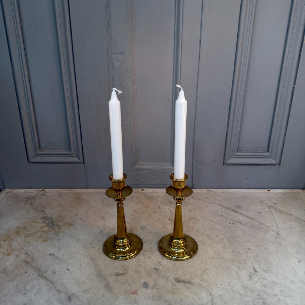 A pair of Arts and Crafts brass candlesticks featuring tapered stems, circular drip pans, and stepped circular bases holding white taper candles. View II
