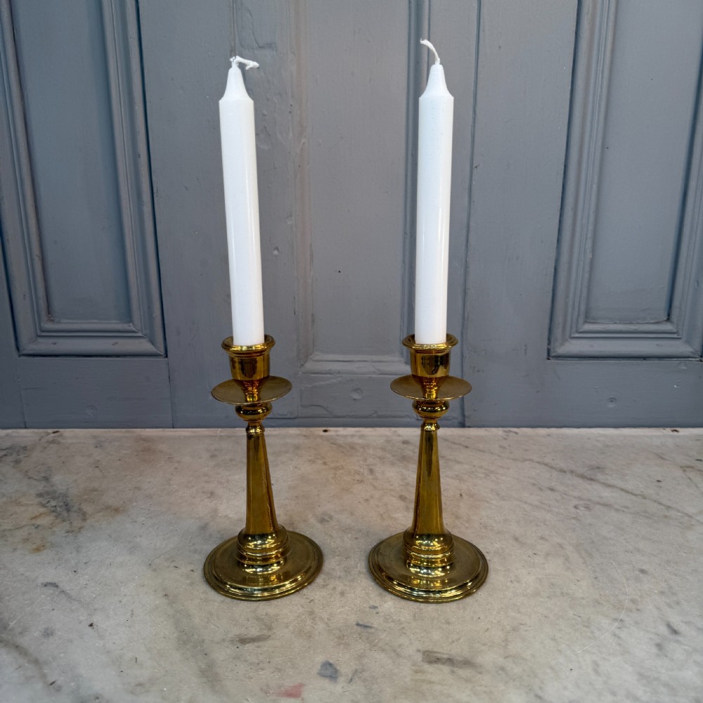 A pair of Arts and Crafts brass candlesticks featuring tapered stems, circular drip pans, and stepped circular bases holding white taper candles. View III