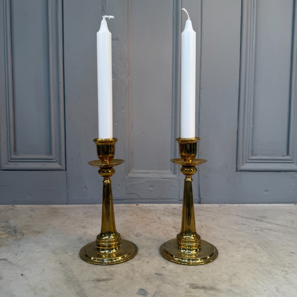 A pair of Arts and Crafts brass candlesticks featuring tapered stems, circular drip pans, and stepped circular bases holding white taper candles. View IV