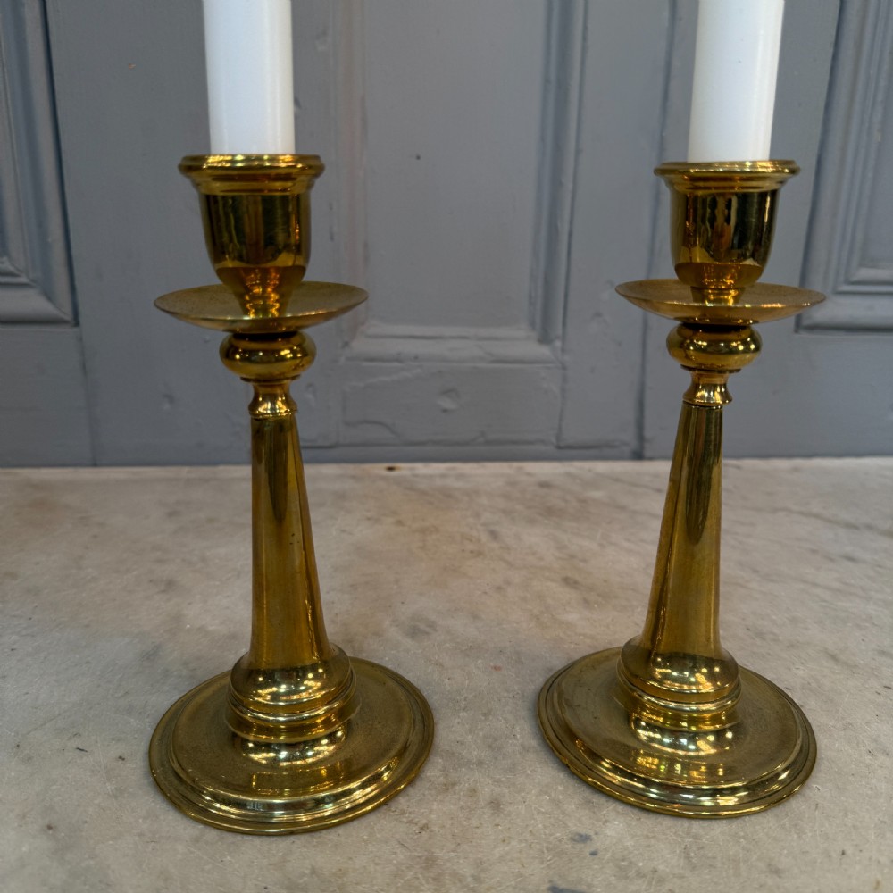 A pair of Arts and Crafts brass candlesticks featuring tapered stems, circular drip pans, and stepped circular bases holding white taper candles. View V