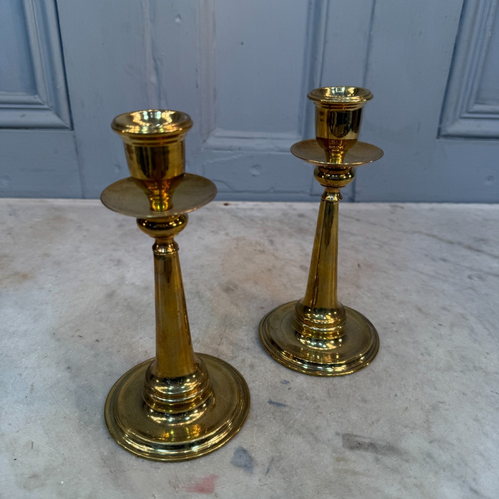 A pair of Arts and Crafts brass candlesticks featuring tapered stems, circular drip pans, and stepped circular bases holding white taper candles. View VI