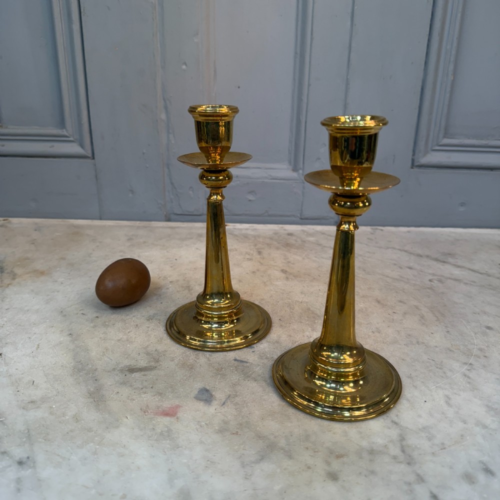 A pair of Arts and Crafts brass candlesticks featuring tapered stems, circular drip pans, and stepped circular bases holding white taper candles. View IX