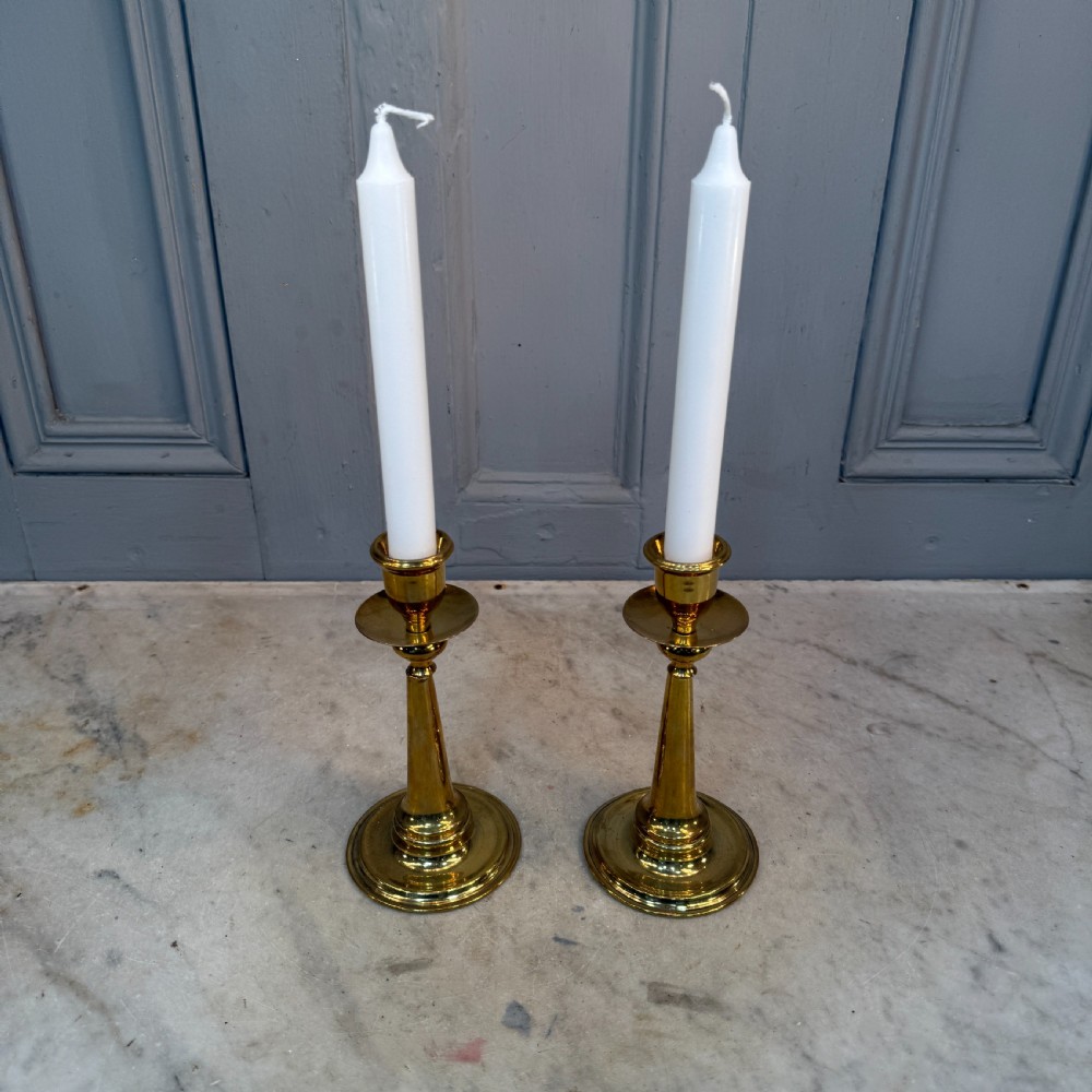 A pair of Arts and Crafts brass candlesticks featuring tapered stems, circular drip pans, and stepped circular bases holding white taper candles. View X