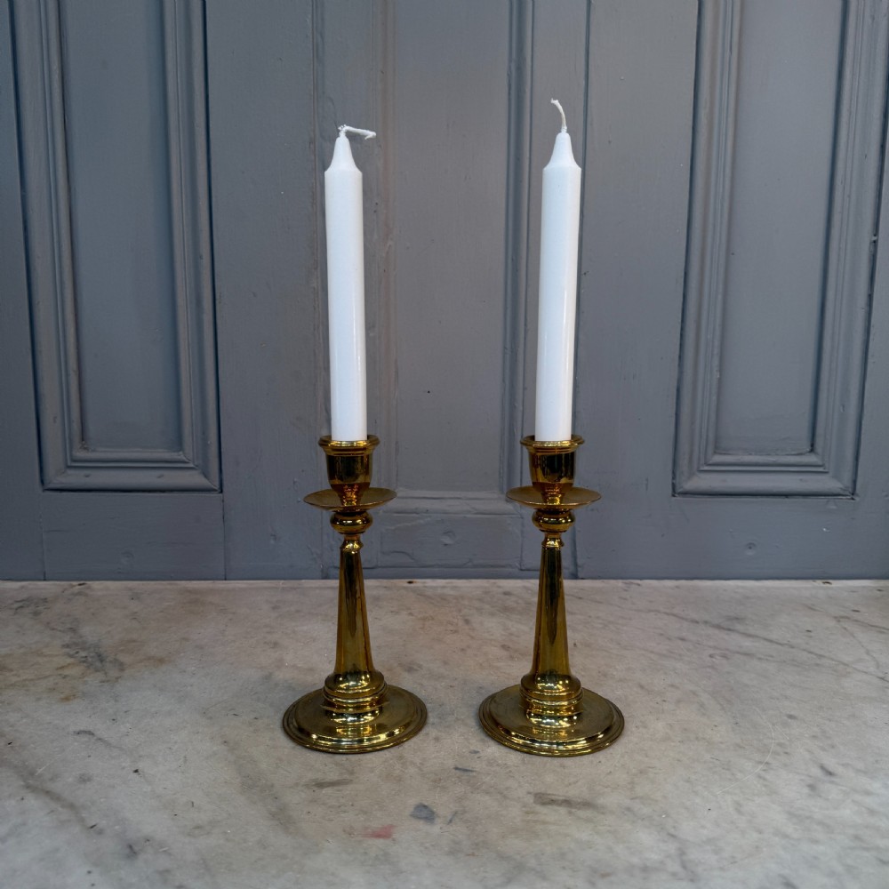 A pair of Arts and Crafts brass candlesticks featuring tapered stems, circular drip pans, and stepped circular bases holding white taper candles. View XI
