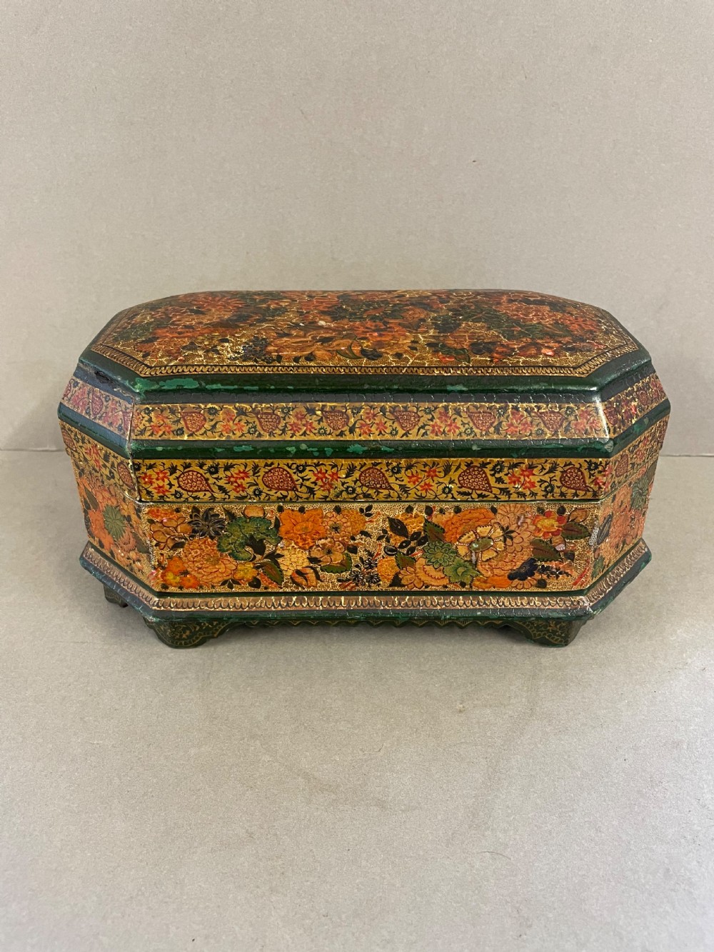 An antique octagonal papier-mache casket featuring intricate hand-painted floral and paisley patterns in green, gold, orange, and black tones.