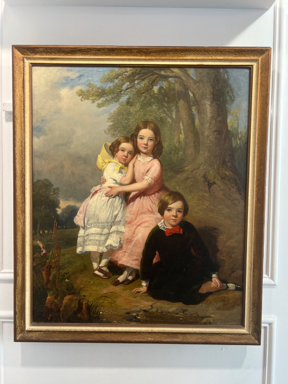 An antique oil painting depicting three young children posed outdoors near a large tree within a molded wooden frame.