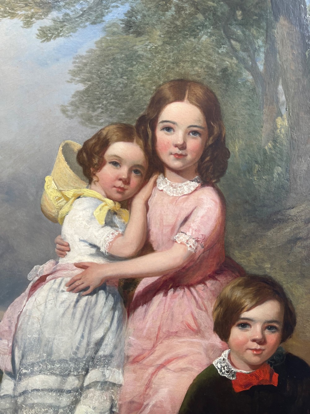 An antique oil painting depicting three young children posed outdoors near a large tree within a molded wooden frame. View II