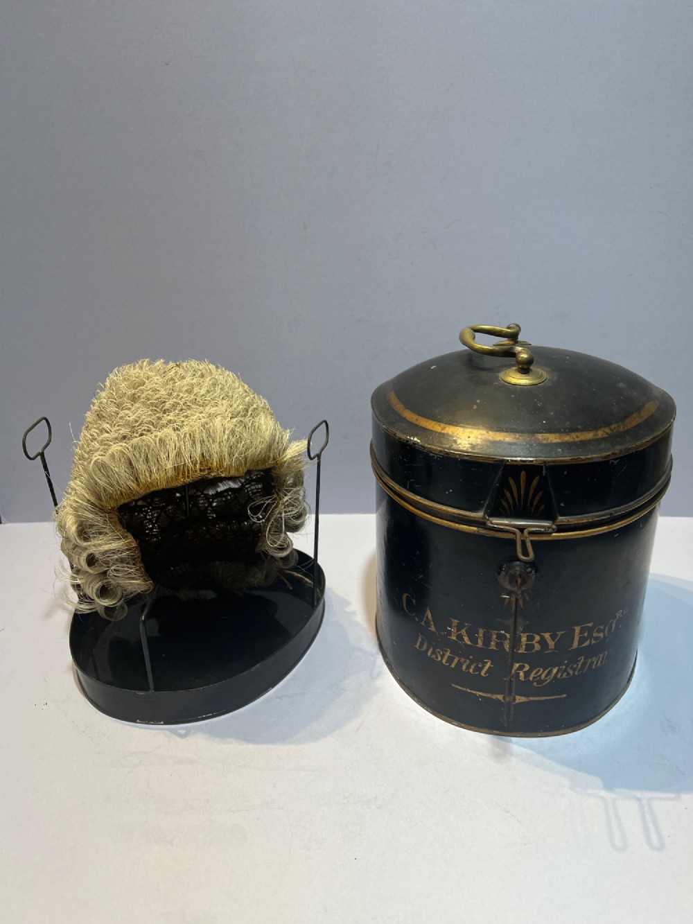 An antique horsehair barrister wig displayed on an internal wire stand next to a black painted metal tin case featuring gold lettering.