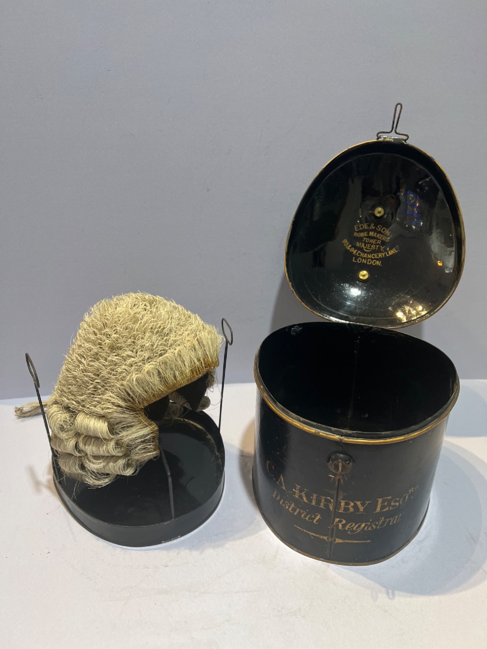 An antique horsehair barrister wig displayed on an internal wire stand next to a black painted metal tin case featuring gold lettering. View II