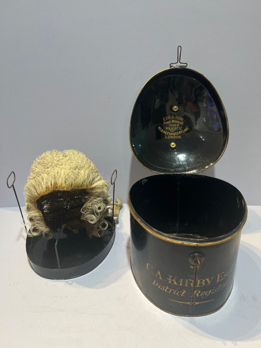 An antique horsehair barrister wig displayed on an internal wire stand next to a black painted metal tin case featuring gold lettering. View III