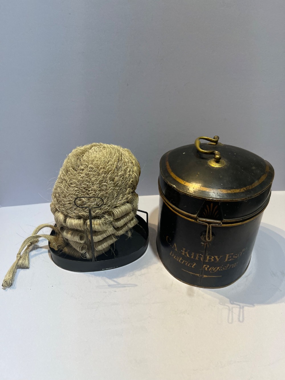An antique horsehair barrister wig displayed on an internal wire stand next to a black painted metal tin case featuring gold lettering. View IV