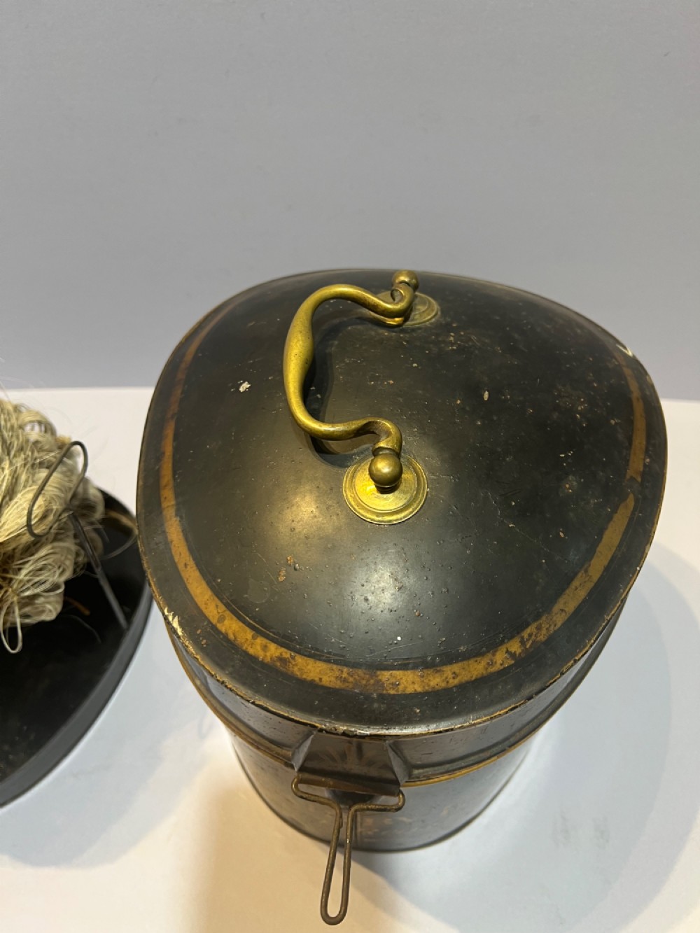 An antique horsehair barrister wig displayed on an internal wire stand next to a black painted metal tin case featuring gold lettering. View V