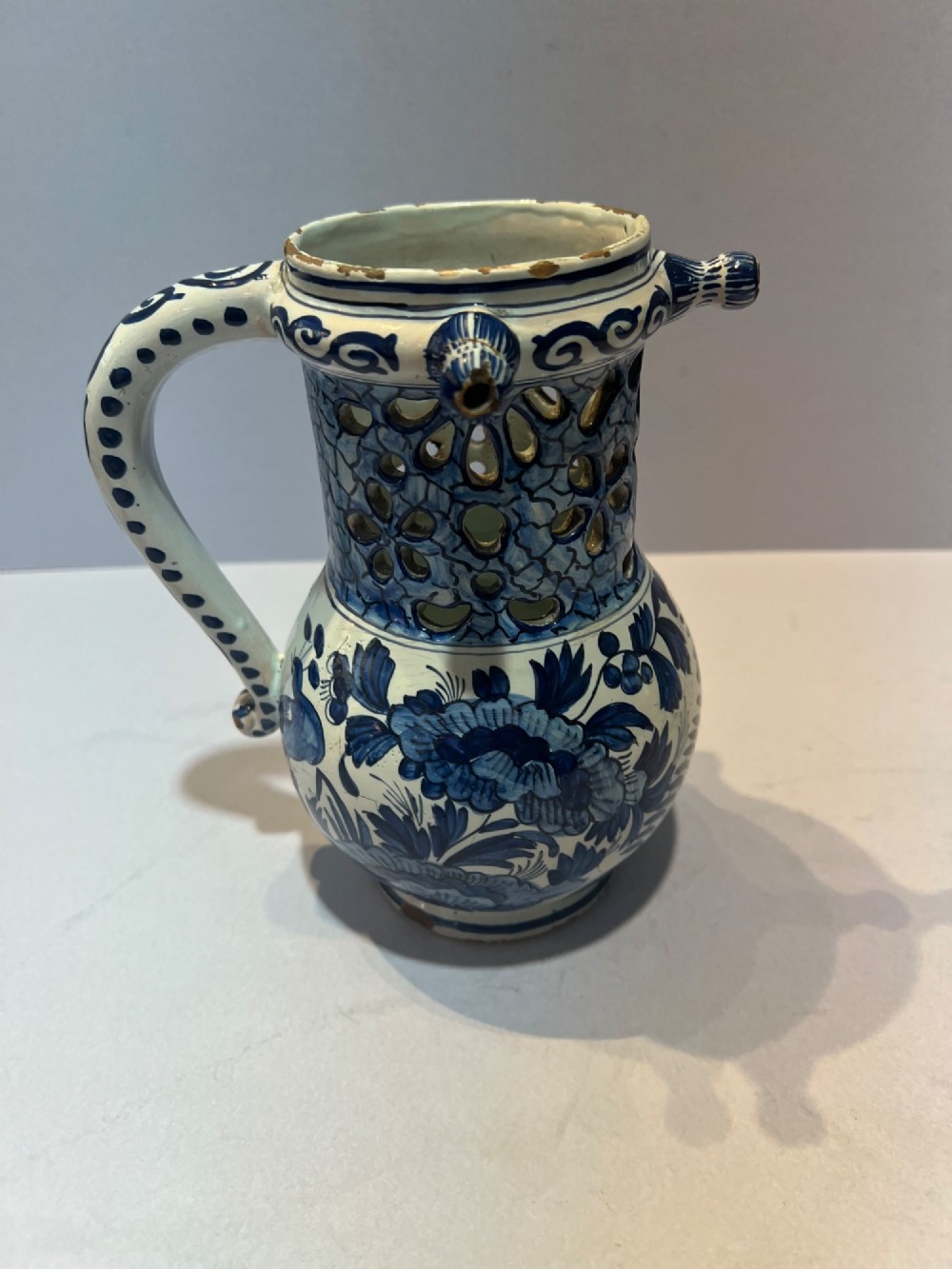 An 18th-century Dutch Delft tin-glazed earthenware puzzle jug featuring hand-painted blue floral motifs, a reticulated neck, and circular spouts along the upper rim.