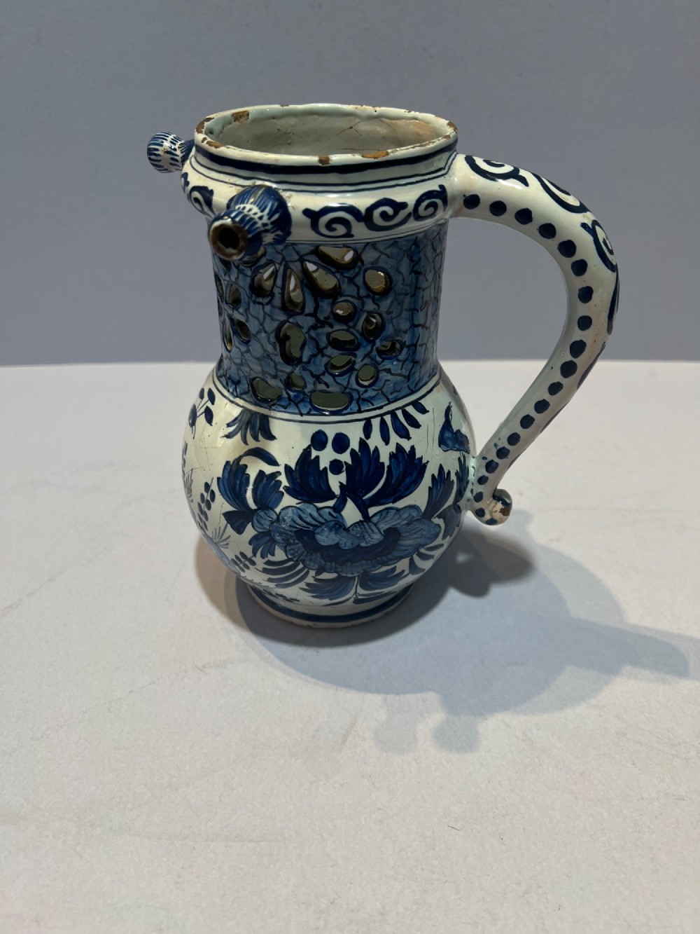 An 18th-century Dutch Delft tin-glazed earthenware puzzle jug featuring hand-painted blue floral motifs, a reticulated neck, and circular spouts along the upper rim. View II