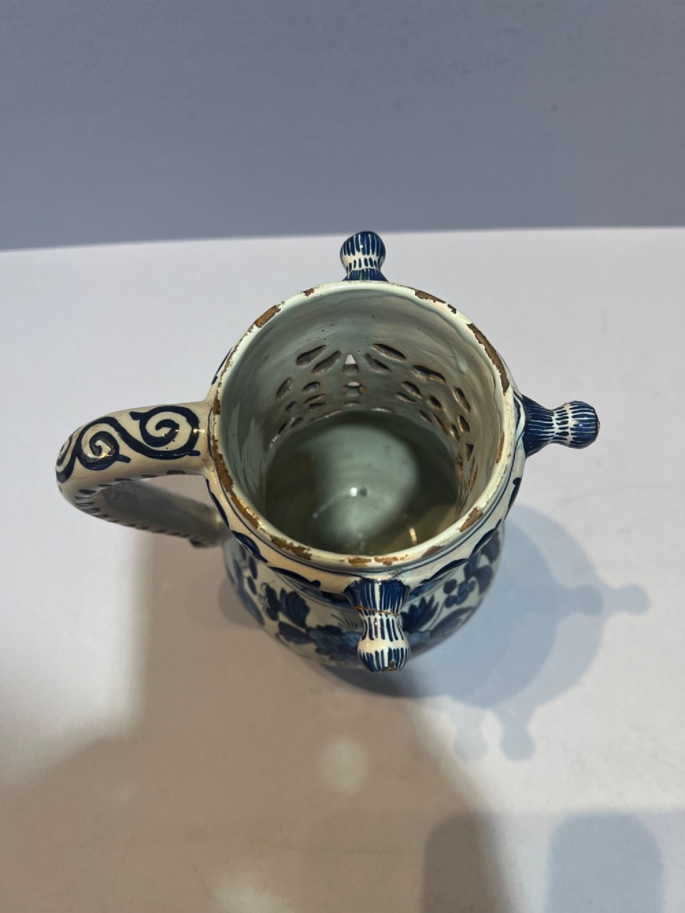 An 18th-century Dutch Delft tin-glazed earthenware puzzle jug featuring hand-painted blue floral motifs, a reticulated neck, and circular spouts along the upper rim. View IV