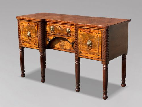 fig 1. A 19th-century George IV flame mahogany breakfront sideboard featuring ebony stringing, lion-mask brass handles, spiral pilasters, and fluted legs.