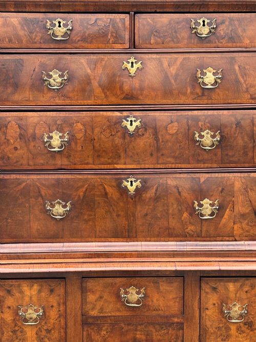 fig 2. An 18th-century Queen Anne walnut chest on stand featuring figured veneers, brass drop handles, a shaped lower apron, and turned legs joined by stretchers.