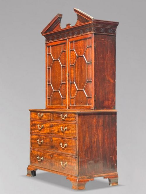 fig 1. An 18th-century George III mahogany bookcase on chest featuring a broken pediment, carved frieze, geometric astragal glazed doors, and four lower drawers with brass handles.