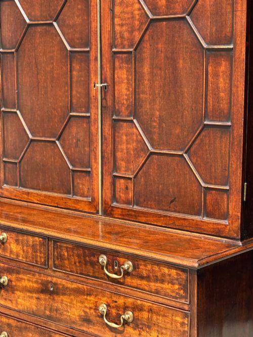 fig 3. An 18th-century George III mahogany bookcase on chest featuring a broken pediment, carved frieze, geometric astragal glazed doors, and four lower drawers with brass handles.