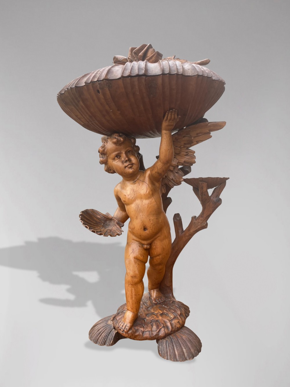 A 19th-century carved wooden figure of a winged putto holding a large scallop shell basin overhead while standing on a shell-shaped base.