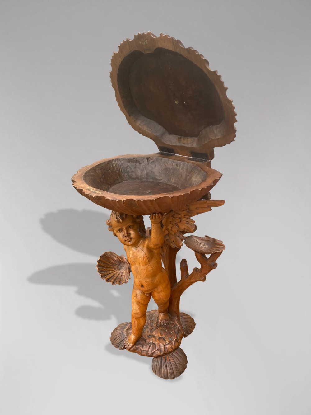 A 19th-century carved wooden figure of a winged putto holding a large scallop shell basin overhead while standing on a shell-shaped base. View III
