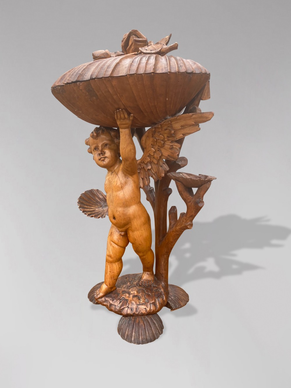 A 19th-century carved wooden figure of a winged putto holding a large scallop shell basin overhead while standing on a shell-shaped base. View IV