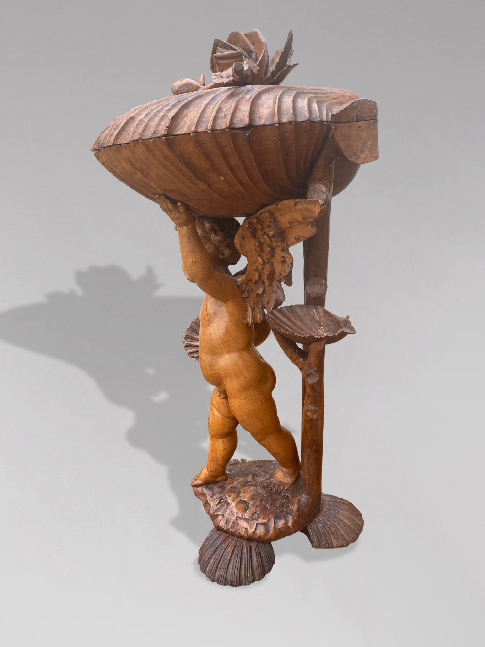 A 19th-century carved wooden figure of a winged putto holding a large scallop shell basin overhead while standing on a shell-shaped base. View V