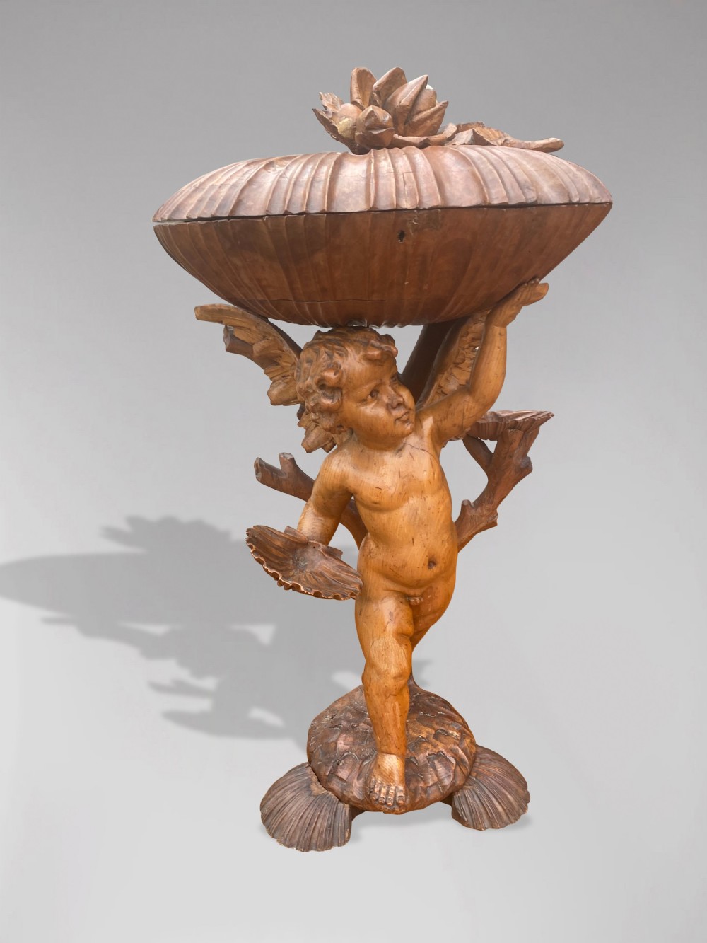A 19th-century carved wooden figure of a winged putto holding a large scallop shell basin overhead while standing on a shell-shaped base. View VIII