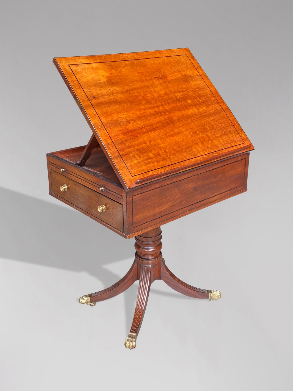 A George III mahogany reading table featuring an adjustable tilted top, a side drawer, a turned pedestal stem, and reeded tripod legs with brass paw feet.
