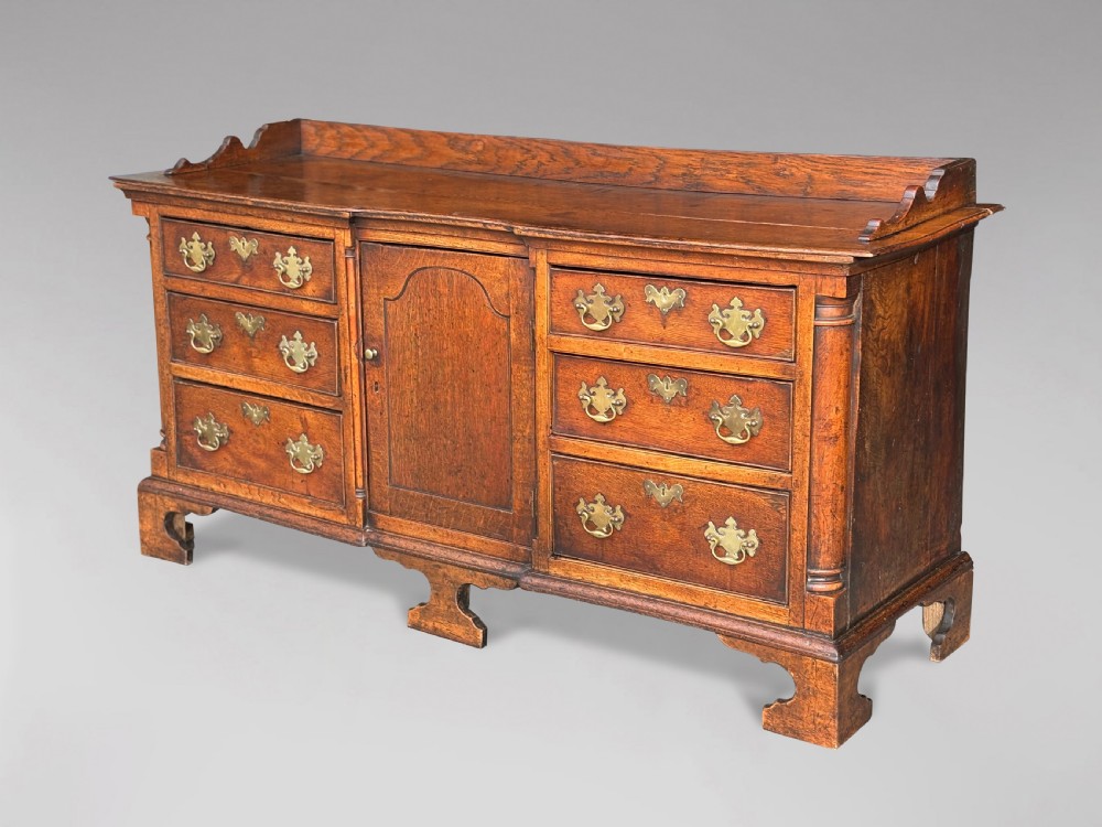 An 18th-century Lancashire oak dresser base featuring brass handles, a central cupboard door, six drawers, and shaped bracket feet.