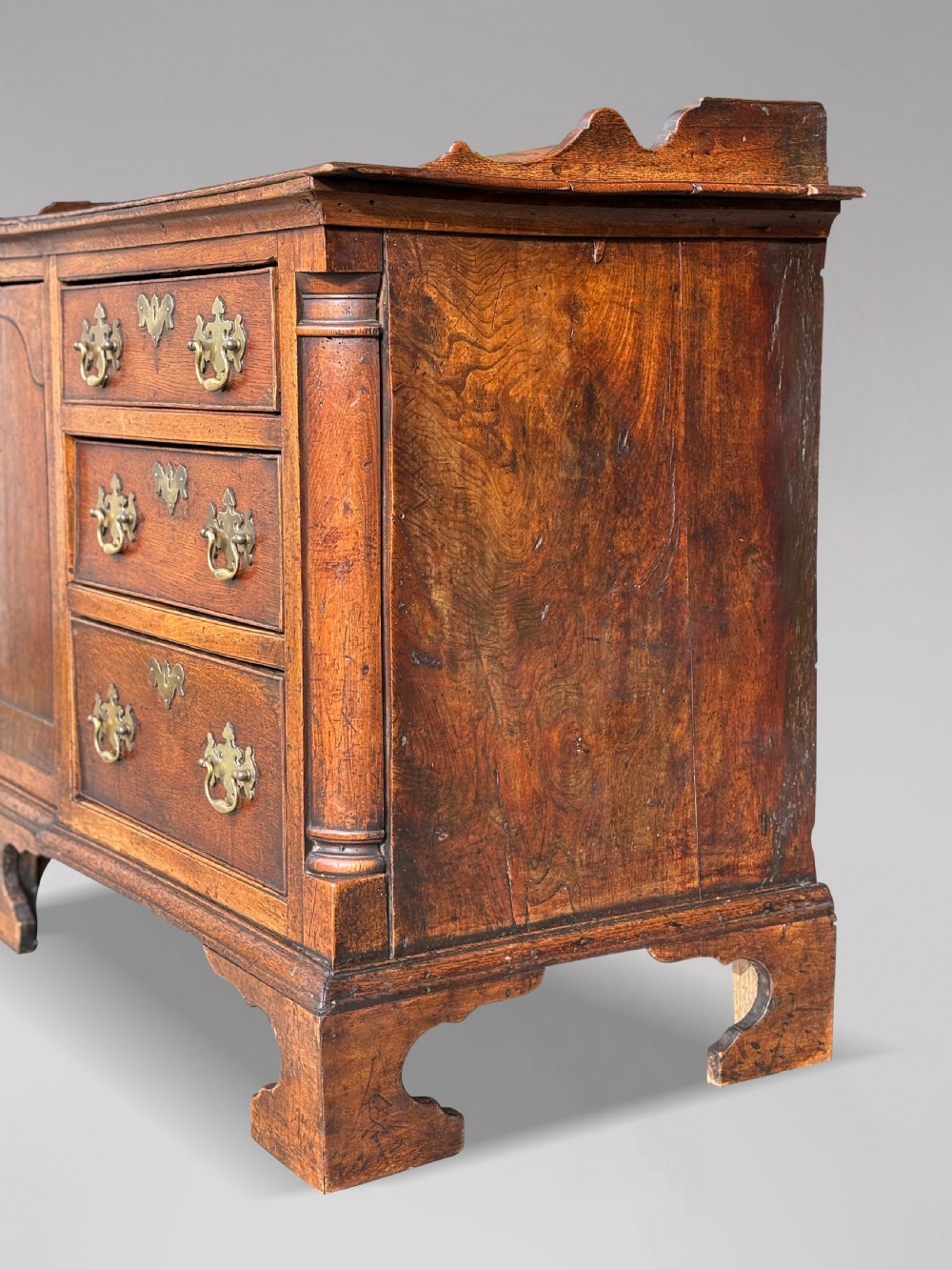 An 18th-century Lancashire oak dresser base featuring brass handles, a central cupboard door, six drawers, and shaped bracket feet. View III