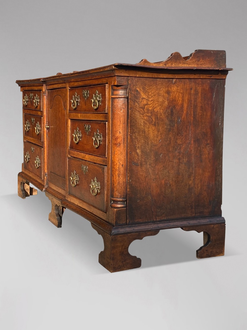 An 18th-century Lancashire oak dresser base featuring brass handles, a central cupboard door, six drawers, and shaped bracket feet. View VII
