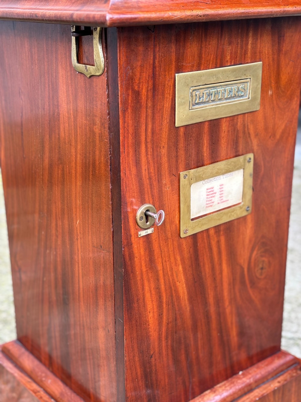 A 19th-century mahogany post box with a molded lid, brass letter slot, collection times plaque frame, and front key lock. View V