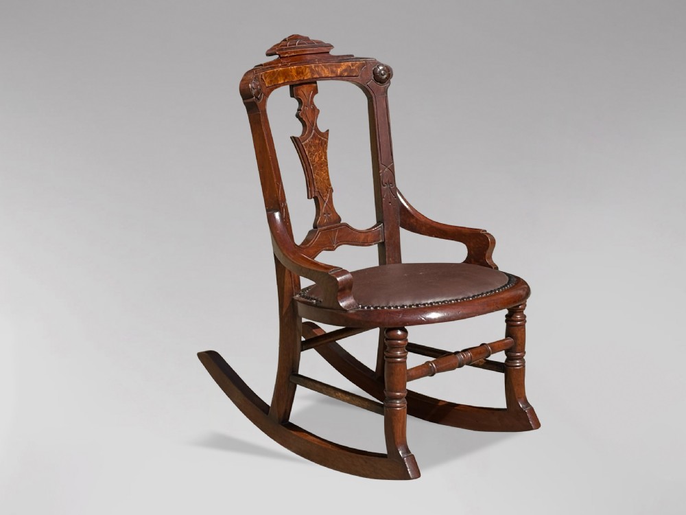 A 19th-century Victorian childs wooden rocking chair featuring a carved backrest with burr wood inlay, a brown leather padded seat with brass stud details, and turned front legs on curved rockers.