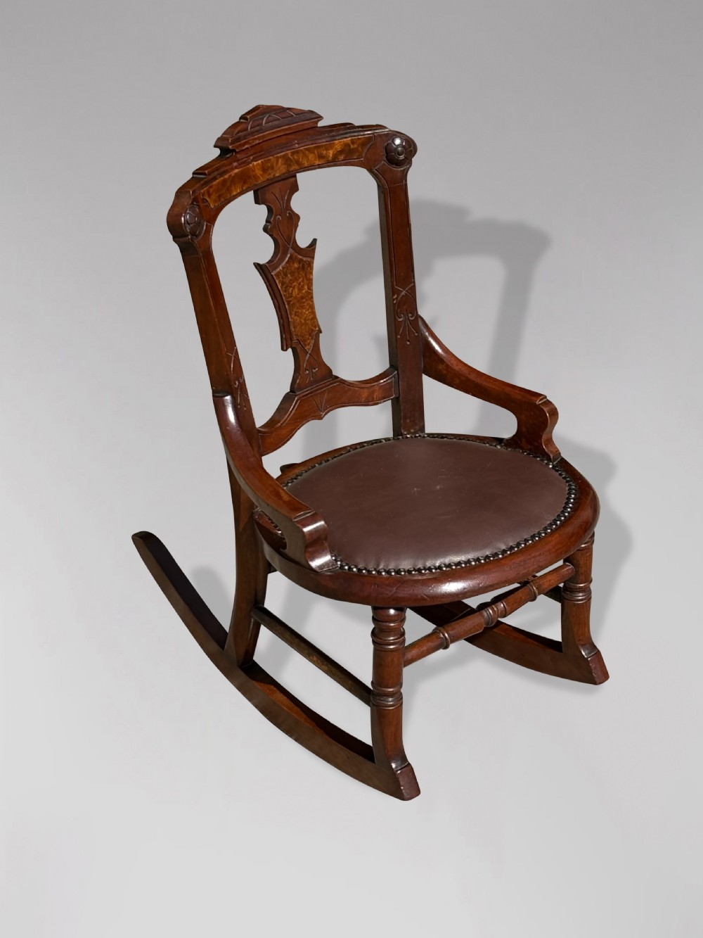 A 19th-century Victorian childs wooden rocking chair featuring a carved backrest with burr wood inlay, a brown leather padded seat with brass stud details, and turned front legs on curved rockers. View II