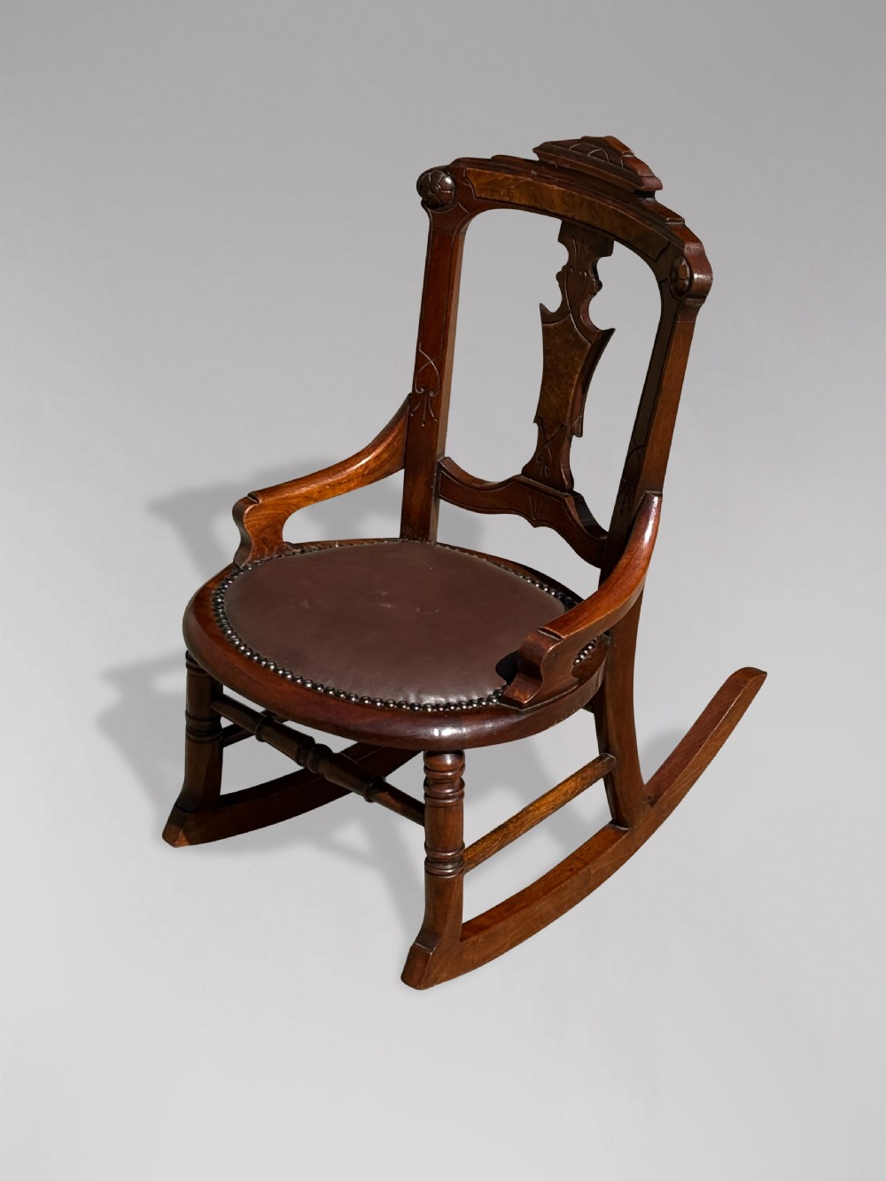 A 19th-century Victorian childs wooden rocking chair featuring a carved backrest with burr wood inlay, a brown leather padded seat with brass stud details, and turned front legs on curved rockers. View IV
