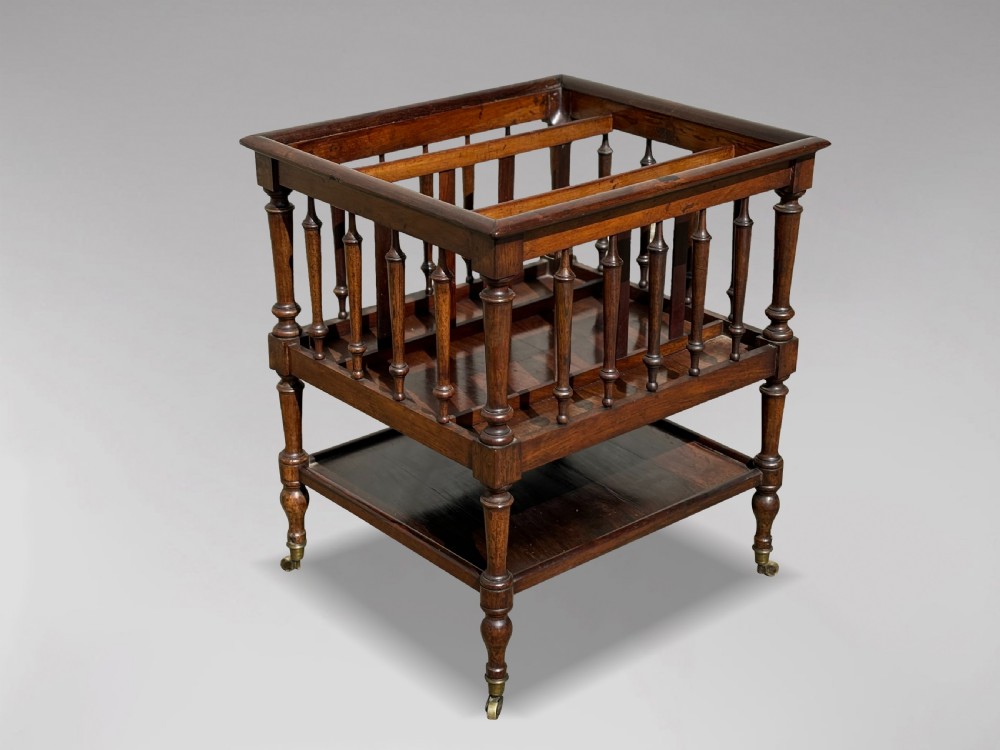 A nineteenth-century dark rosewood canterbury featuring turned spindle supports, divided upper compartments, a lower open shelf, and brass castors.