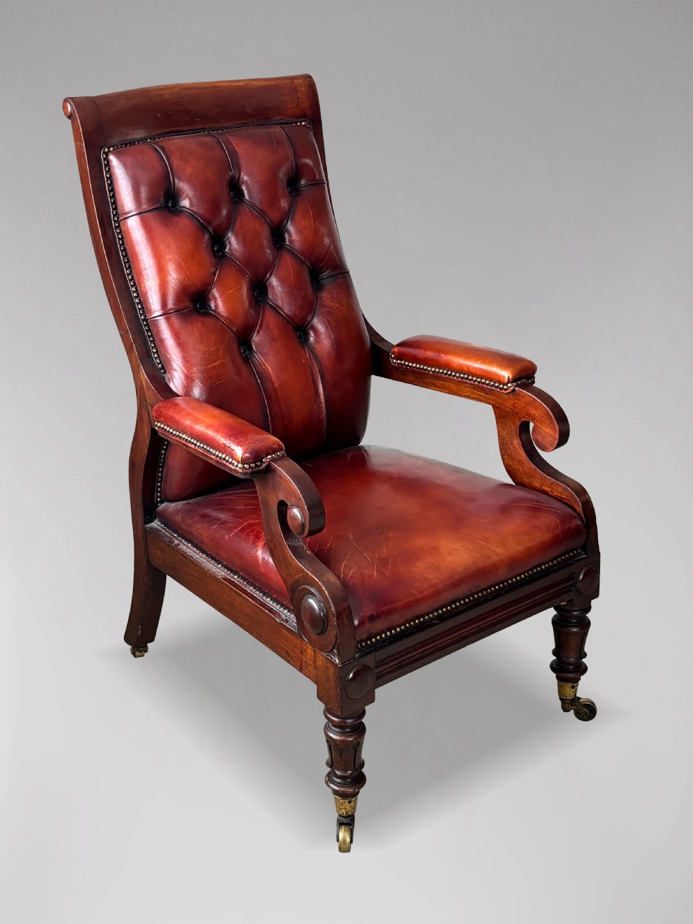 A 19th-century William IV mahogany library armchair featuring deep-buttoned red-brown leather upholstery, scrolled arms, brass stud trim, and turned front legs on brass castors.