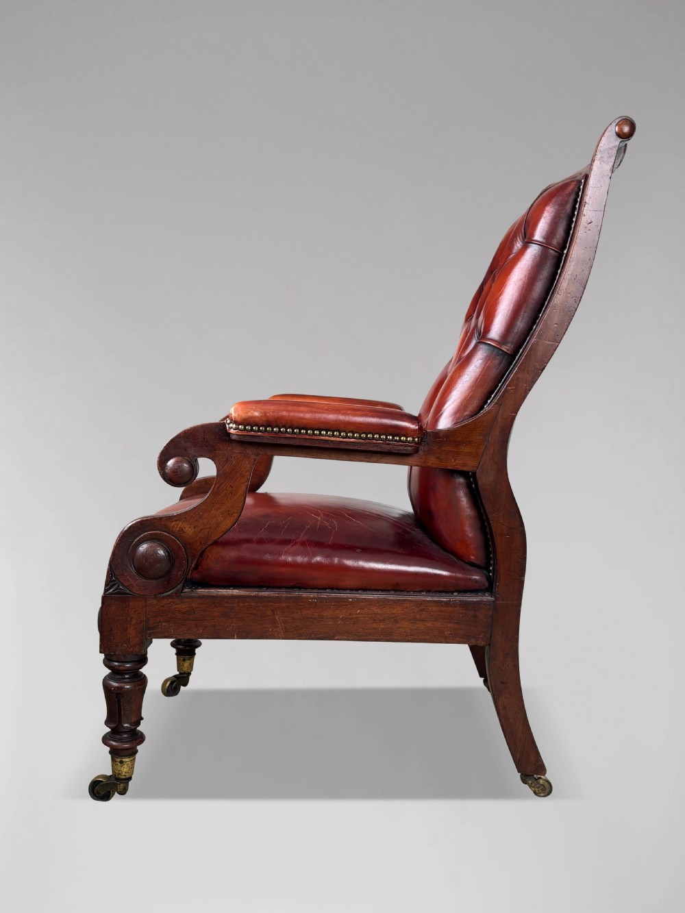 A 19th-century William IV mahogany library armchair featuring deep-buttoned red-brown leather upholstery, scrolled arms, brass stud trim, and turned front legs on brass castors. View III